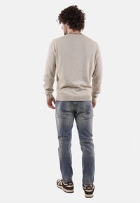 Maglione beige con un design testurizzato, polsini e orlo a coste. Indossato con jeans chiari sdruciti e sneakers marroni con accenti bianchi.
