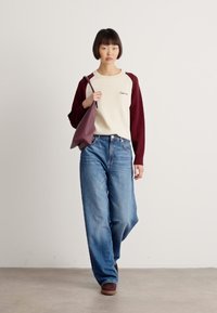 Young person walking indoors wearing a cream and burgundy sweatshirt, wide-leg blue jeans, brown shoes, and carrying a burgundy shoulder bag.