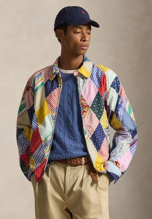 Nuori mies, jolla on värikäs tilkkutäkki-takki, sininen palmikkoneulepusero, beiget housut, punottu vyö ja laivastonsininen lippalakki, katse sivulle.