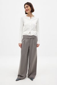 Cárdigan blanco de punto con cuello redondo y botones, combinado con pantalones gris holgados que presentan pliegues y una cintura plana. Zapatos grises de punta.