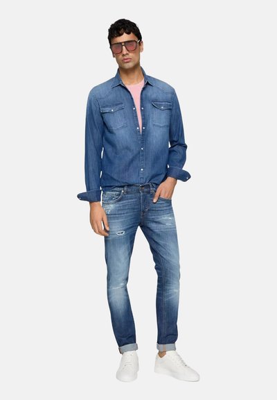 Camicia di denim in blu medio con tasche sul petto, maniche lunghe arrotolate, abbinata a jeans chiari strappati e sneaker bianche. T-shirt rosa sotto.