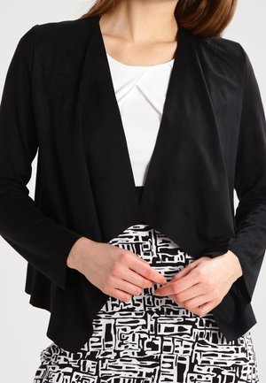 Kvinde iført en sort åbent front-jakke over en hvid top med et geometrisk sort-hvidt mønstret skørt, hænderne samlet ved livet.