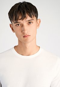 T-shirt bianca con scollo a girocollo in cotone morbido, caratterizzata da un logo discreto sul petto, indossata da una modella con capelli scuri e sfondo neutro.