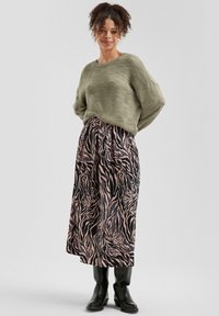 Olivgrüner Strickpullover kombiniert mit einem langen, schwarzen Rock mit einem pinken Tigerstreifenmuster und schwarzen knielangen Stiefeln.