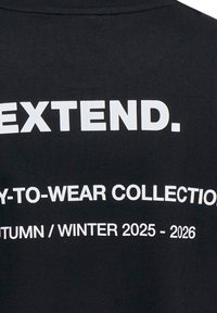 T-shirt nera con testo bianco in grassetto: "EXTEND. COLLEZIONE PRONTA DA INDOSSARE AUTUNNO/INVERNO 2025-2026" sul retro. Tessuto di cotone liscio.
