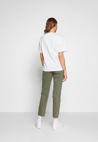 Weißes Baumwoll-T-Shirt mit kurzen Ärmeln, kombiniert mit olivgrünen Cordhosen und weißen Sneakern. Modell mit dem Rücken zur Kamera.