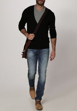 Mann geht und trägt eine schwarze Strickjacke über einem grauen Hemd, blaue Jeans, hellbraune Schuhe und eine braune Umhängetasche.