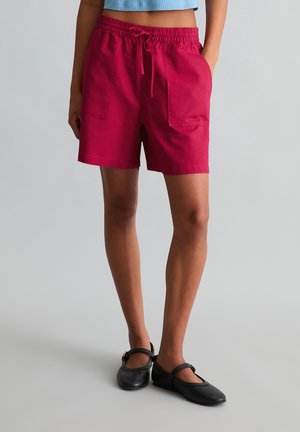 Person trägt knallrote Shorts mit Kordelzug und großen Fronttaschen sowie schwarze Mary-Jane-Schuhe, steht vor einem einfarbigen Hintergrund.