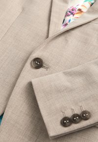 Jasnobeżowa marynarka o delikatnej teksturze, z brązowym guzikiem i trzema dodatkowymi guzikami na mankiecie, wykończona kwiatową podszewką.