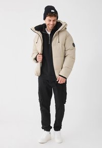 Beżowa kurtka puchowa z kapturem, czarną podszewką, zapinana na zamek, oraz czarne spodnie cargo. Dodatki to granatowa czapka beanie i białe trampki.