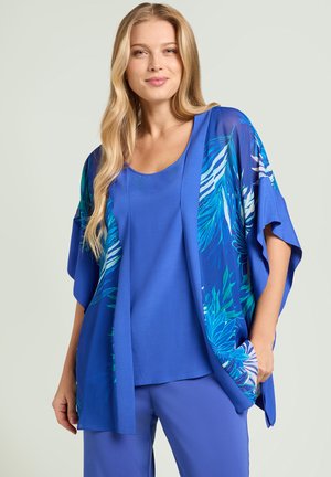 Kobieta nosząca niebieski strój z topem bez rękawów i pasującą kimonową kurtką z turkusowo-białymi wzorami liści.