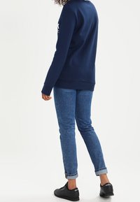 Marinblå sweatshirt med ribbade muddarna, i kombination med uppvikta blå jeans och svarta sneakers, sedd bakifrån.
