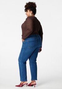 Bruin getextureerd shirt met lange mouwen gecombineerd met blauwe jeans met rechte pijpen. Heeft achterzakken en rode schoenen met hak. Witte achtergrond.