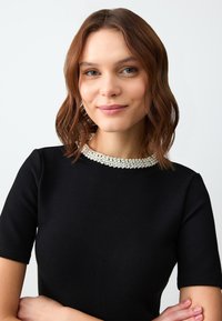 Czarna sukienka z gładkiego materiału z okrągłym, teksturowanym dekoltem zdobionym ornamentowymi akcentami przypominającymi perły. Krótkie rękawy; dopasowany krój.