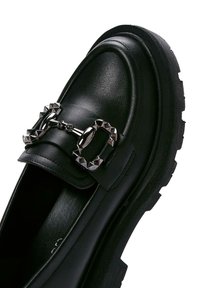 Mocassins en cuir noir avec une semelle épaisse, des clous décoratifs en métal argenté et un détail de boucle saisissant sur le dessus.