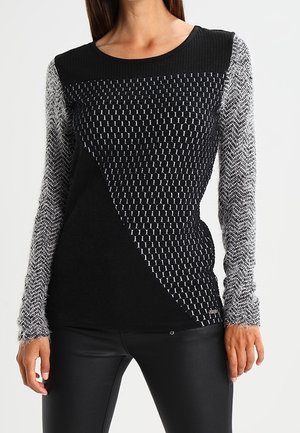 Femme portant un haut en maille noir et gris à manches longues avec des motifs géométriques et en chevrons, associé à un pantalon en cuir noir.