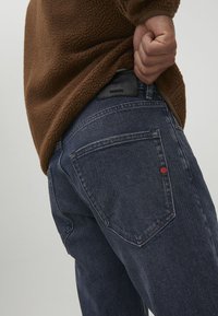 Jeans de mezclilla azul oscuro con un acabado texturizado; presentan un bolsillo de parche y un pequeño logotipo rojo en la parte posterior, combinados con una parte superior de forro polar marrón.