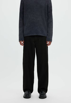 Personne portant un pull côtelé foncé, un pantalon noir ample et des chaussures noires, debout devant un fond clair uni.