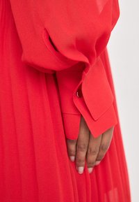 Crvena haljina labavog kroja s dugim rukavima; ima dugmad na manžetama i tekući, teksturirani materijal. Boja je svijetla i ujednačena.