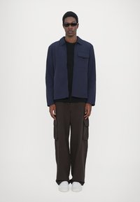 Navy jacket with a front pocket over a black shirt, paired with brown wide-leg cargo pants and white sneakers. Smooth texture, tailored fit.