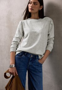 Felpa grigia chiara con maniche a sbuffo e sottili decorazioni, abbinata a jeans in denim blu. Il modello tiene in mano una borsa marrone e un bracciale d'argento.