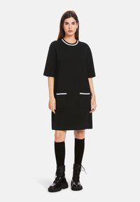 Schwarzes Strickkleid mit kurzen Ärmeln, das einen weißen Besatz am Ausschnitt und zwei Fronttaschen hat. Getragen mit schwarzen knielangen Socken und Stiefeln.