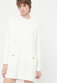 Persoană tânără îmbrăcată într-o rochie albă din tricot cu modele împletite, cu mâneci lungi și două buzunare frontale cu butoane aurii, stând pe un fundal alb.
