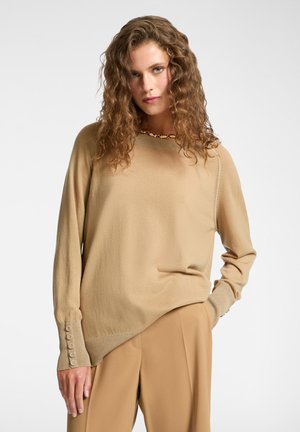 Una mujer con cabello rizado lleva un suéter largo de manga larga beige con botones en los puños y pantalones beige a juego, posando frente a un fondo liso.
