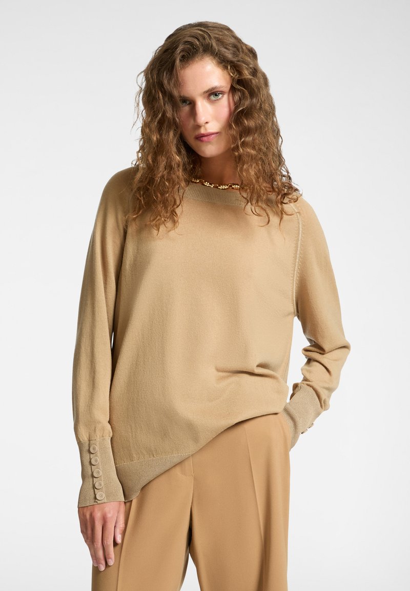 Donna con capelli ricci indossa un maglione beige a maniche lunghe con bottoni sui polsini e pantaloni beige coordinati, in posa davanti a uno sfondo semplice.