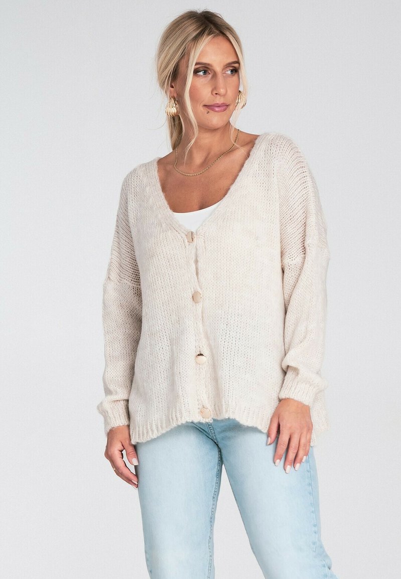 Beżowy sweter z dzianiny z dekoltem w kształcie V, trzema guzikami z przodu i ściągaczami na rękawach, w połączeniu z jasnoniebieskimi dżinsami.