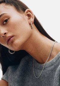 Hõbedast rõngaskõrvarõngad ja peen kett-kaelakee, mõlemad kaunistatud väikeste säravate detailidega, tavalise tausta ees.