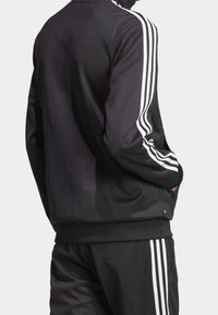 Person i svart träningsoverall med tre vita ränder på ärmarna och byxorna, med händerna i jackfickorna, vänd bort.