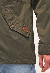 Olivgrön jacka med en texturerad yta, som har en sidficka med tryckknapp och en brunt läderlapp med sömnadsdetaljer.