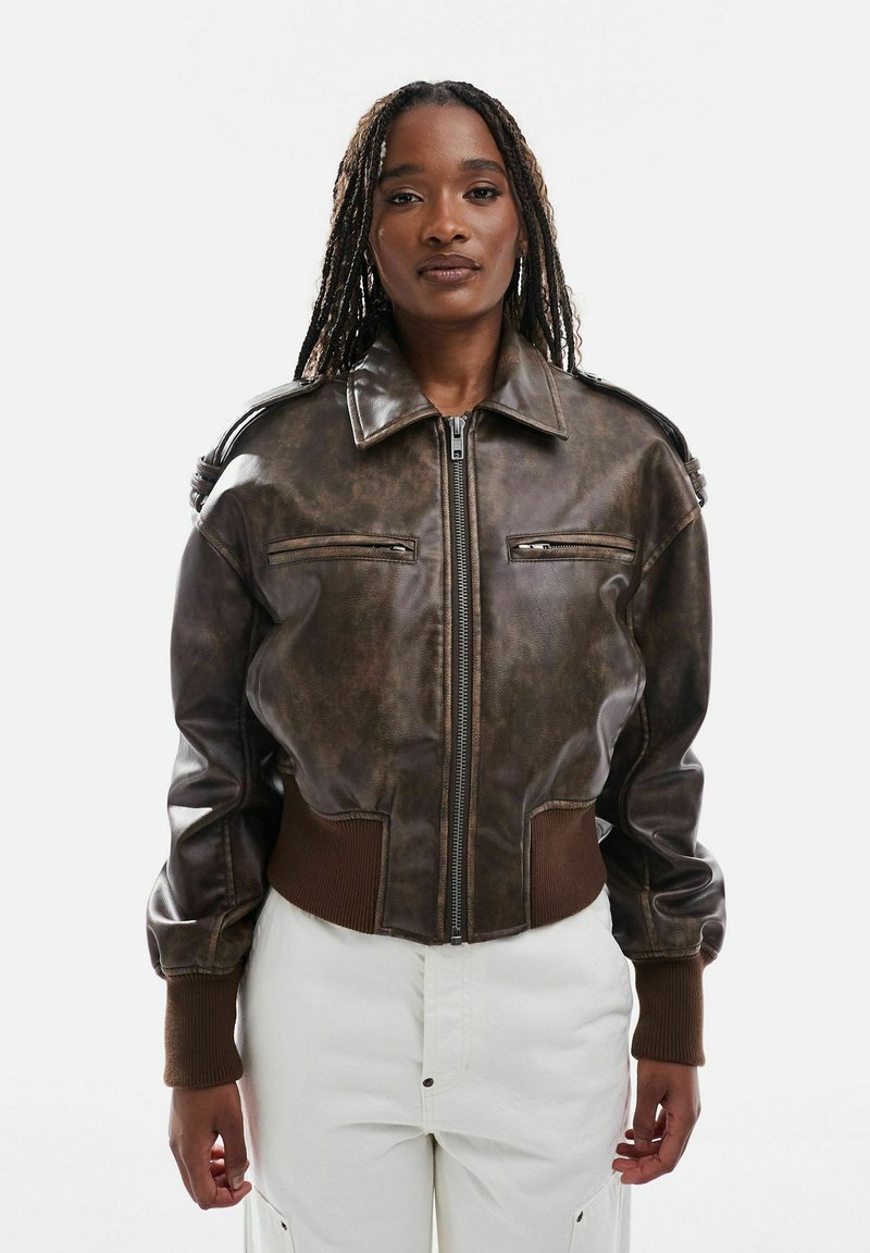 Femme aux longues tresses portant une veste en cuir marron à fermeture éclair avec des poignets côtelés et un pantalon blanc, face à l'avant sur un fond uni.