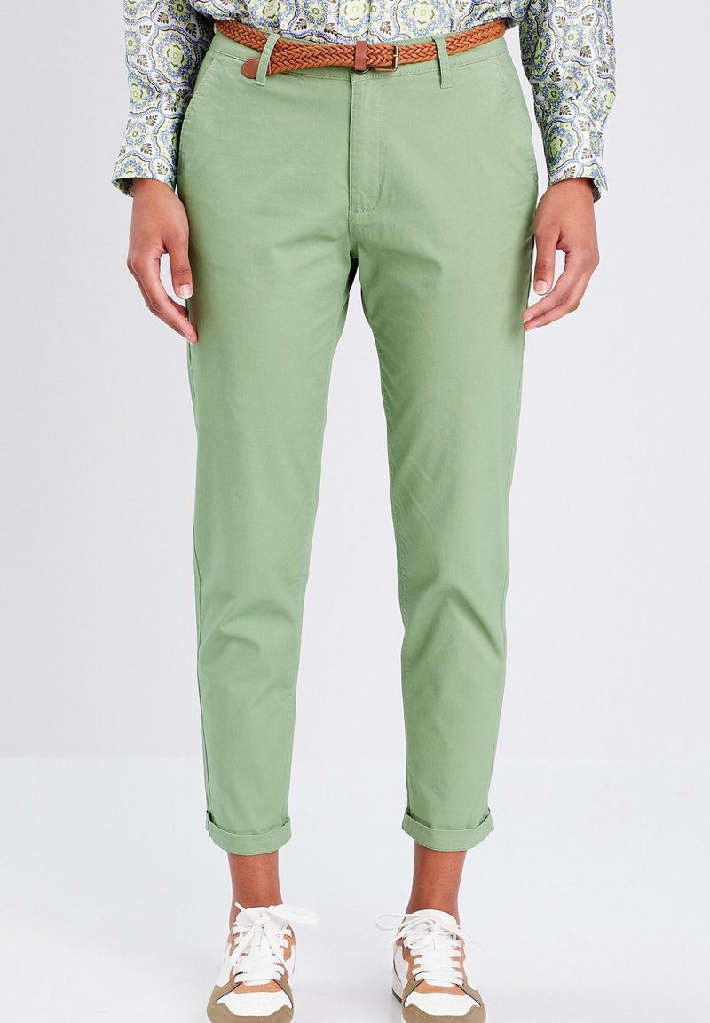 Chinos vert clair en coton, présentant une coupe slim, des ourlets retroussés, et une ceinture tressée marron. Associés à des baskets blanches et beige.