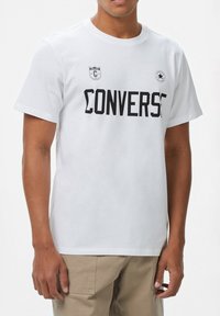 Camiseta de algodón blanca con texto negro "CONVERSE", logotipo en el pecho y mangas cortas. Textura suave con un ajuste estándar.