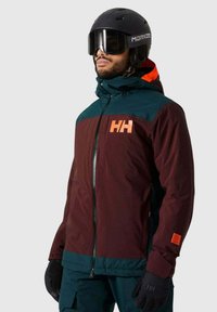 Bordeauxfarbene und petrolfarbene Skijacke mit hohem Kragen und orangefarbenen Akzenten, ausgestattet mit einem Reißverschluss vorne und Logo. Kombiniert mit schwarzen Handschuhen und Helm.