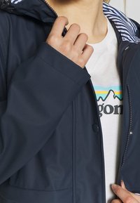 Mörkblå vattentät jacka med huva, som har ett randigt foder, tryckknappar och en slät yta. En grafisk t-shirt syns delvis under.