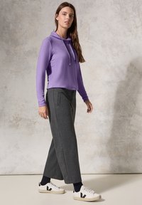 Violetti pitkähihaisen hupparin, jossa on nyörit, yhdistetty harmaisiin löysiin housuihin ja valkoisiin tennareihin, joissa on mustia yksityiskohtia. Sileä materiaali.