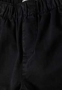Zwarte broek in close-up, met elastische tailleband en zak met zichtbare stikdetails op de tekstuurstof.