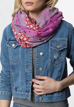 Personne blonde portant une veste en jean bleue et une écharpe à motif d'étoiles violettes et rouges, ajustant la veste d'une main.
