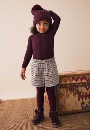 Dziecko ubrane w bordowy dzianinowy sweter, czapkę z pomponem, bordowe rajstopy, wzorzyste spodenki i błyszczące bordowe buty stojące przy zabytkowej skrzyni.