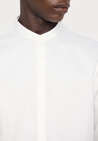 Haut du corps d'une personne portant une chemise blanche avec un col mandarin et des boutons visibles, tête légèrement tournée sur le côté.