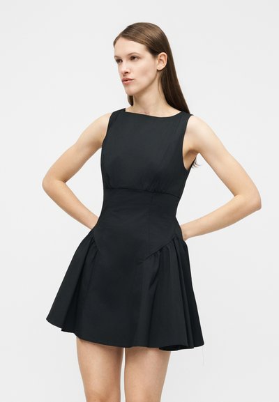 Νεαρή γυναίκα που στέκεται με τα χέρια στη μέση, φορώντας ένα μαύρο εφαρμοστό αμάνικο φόρεμα με φούστα που ανοίγει.