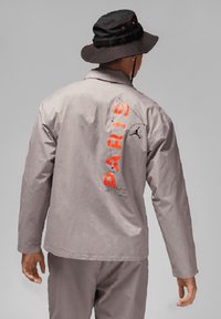Lichtgrijze jas met "PARIS" in neonoranje op de achterkant gedrukt. Met Jordan-logo, een kraag en lange mouwen. Gedragen met een zwarte bucket hat.