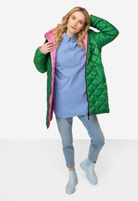 Chaqueta acolchada verde y rosa, camiseta a rayas azul, jeans de un azul claro y botas de goma a juego. La chaqueta cuenta con una capucha y cierre de cremallera.