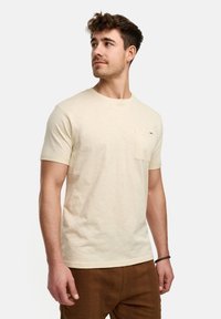 Camiseta beige claro con cuello redondo, mangas cortas y un bolsillo en el pecho izquierdo. La tela parece suave con una sutil textura.