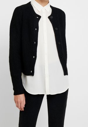 Zwarte gebreide cardigan met tekstuurstof, ronde halslijn en zes zwarte knopen. Gedragen over een witte blouse met kraag.