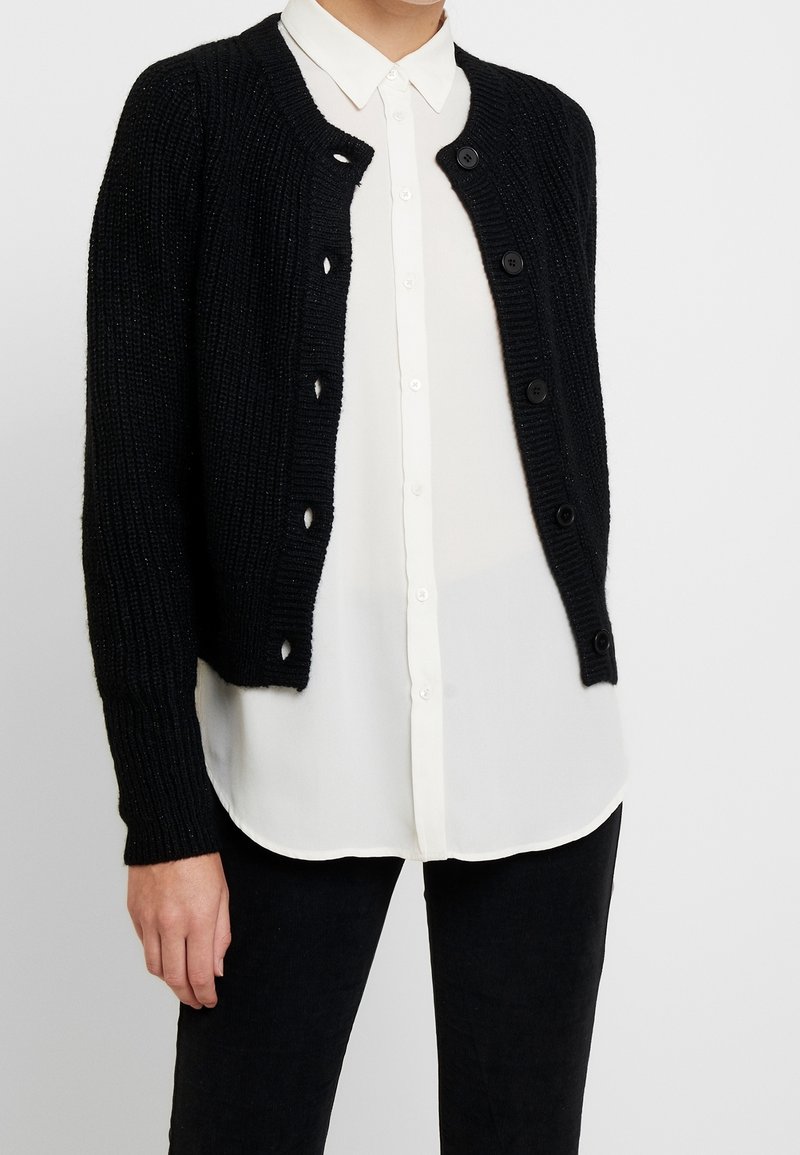 Schwarze Strickjacke aus strukturiertem Stoff, mit rundem Halsausschnitt und sechs schwarzen Knöpfen. Getragen über einem weißen Hemd mit Kragen.