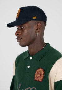 Groene en beige poloshirt met korte mouwen, geborduurd logo in castanjebruin en witte knopen. Donkerblauwe pet met gouden accenten.